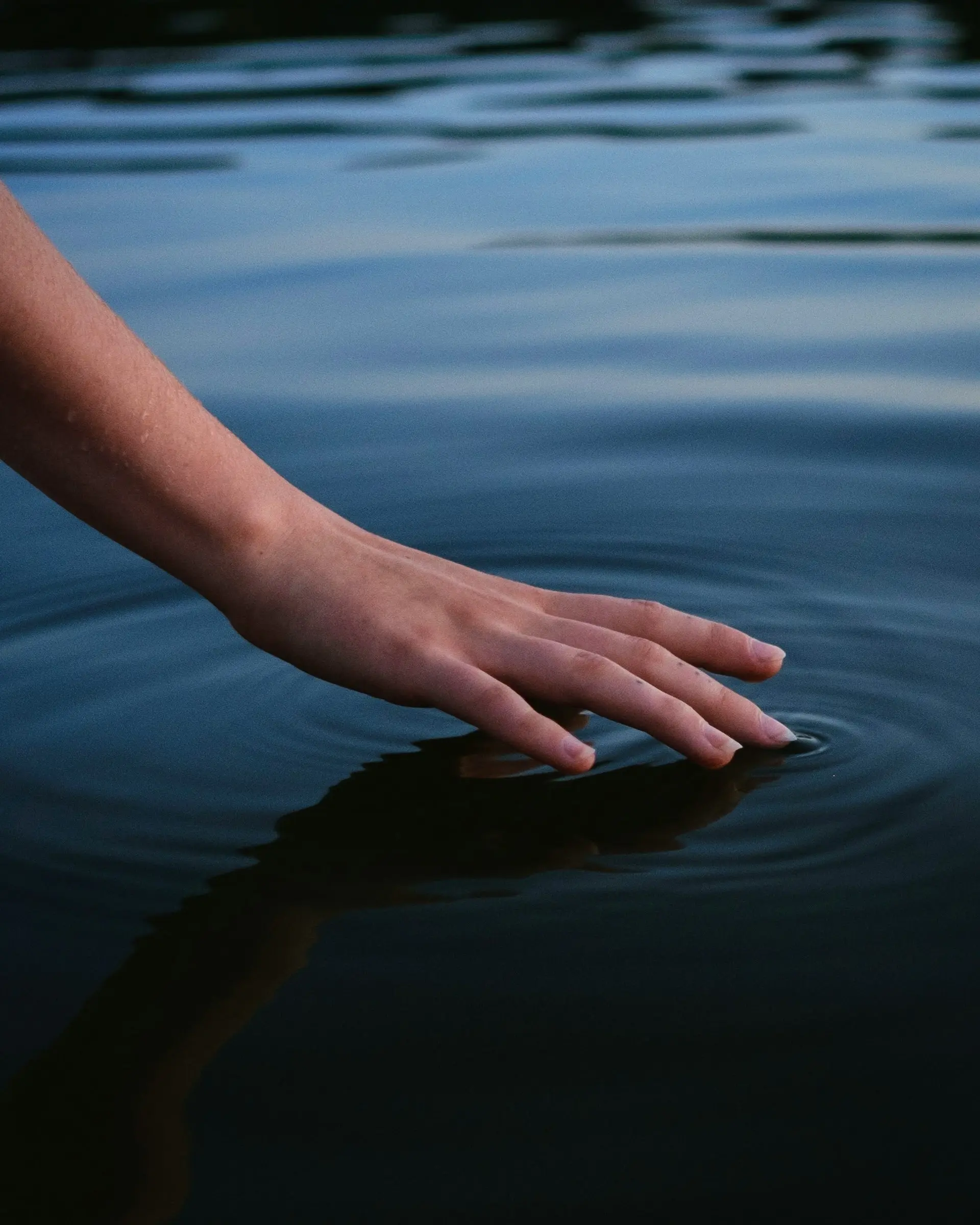 miguel-bandeira-6vHGBZ2A5Rc-unsplash Hand gently touching calm water, creating rippling circles.